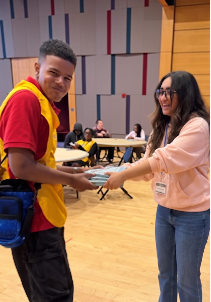 Sophia handing a stack of financial-literacy books to a young man in a red and yellow uniform, with students seated at round tables behind them.